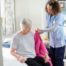 Caregiver gently helping an older woman put on a pink sweater while seated on her bed at home.