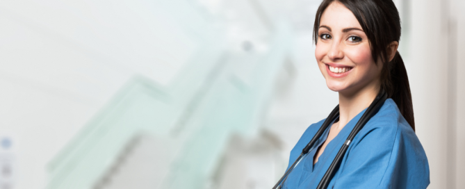 Female caregiver in blue scrubs stands with arms crossed and stethoscope around her neck with a big simile on face in front of staircase in a home care agency.