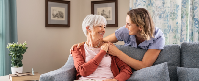 Smiling caregiver with hand over the shoulder of a happy elderly woman sitting on the couch.