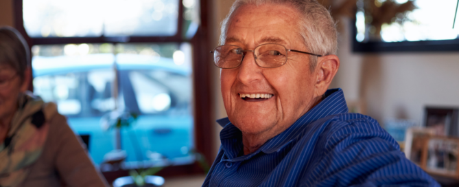 A smiling senior man sitting at a table, happy, as a result of successful long-distance caregiving.