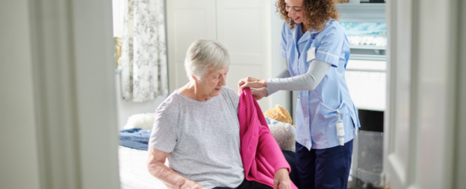 A professional caregiver helps a senior client get her coat on and get ready for the day.