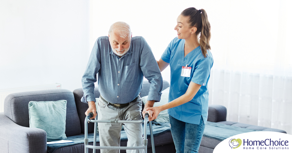 A professional caregiver helps an older man walk with a walker, showing how having a caregiver help a loved one stay mobile safely can help avoid hospital readmissions.
