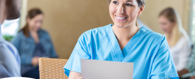 A woman in scrubs holds a paper while interviewing with someone else, representing how a solid caregiver resume can get you to an interview.