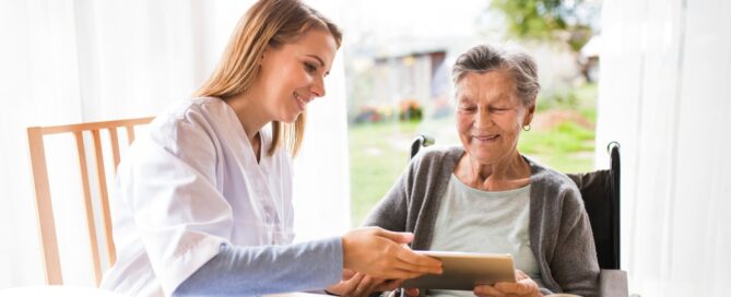 A professional caregiver smiles as she helps a happy senior client with her tablet, showing that she is managing caregiving stress well.