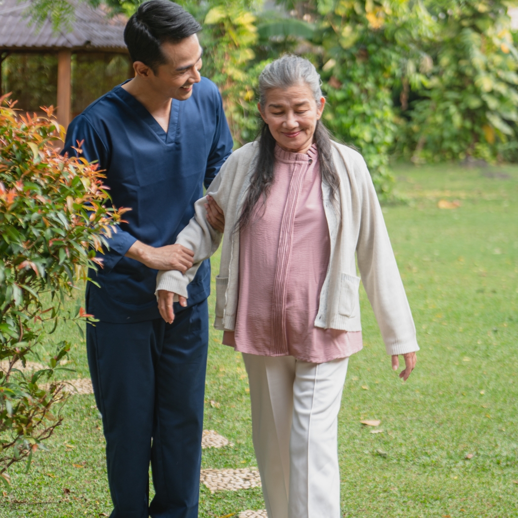 Home Care in Durham (1) A young Asian male caregiver walks with an elderly Asian woman in their garden, highlighting the gentle support and companionship shared through outdoor moments.