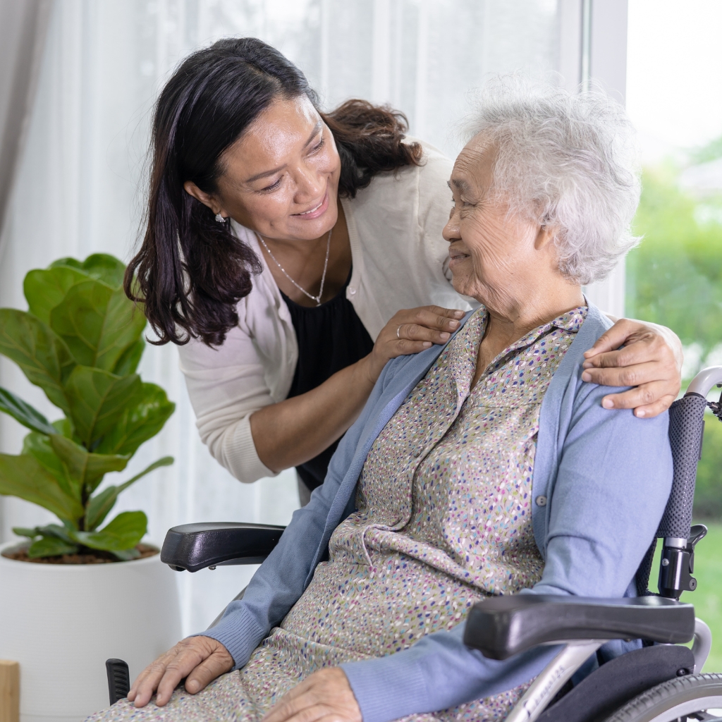 Home Care in Durham An Asian caregiver with black hair looks at an elderly Asian woman in a wheelchair indoors, capturing a quiet moment of care and connection.