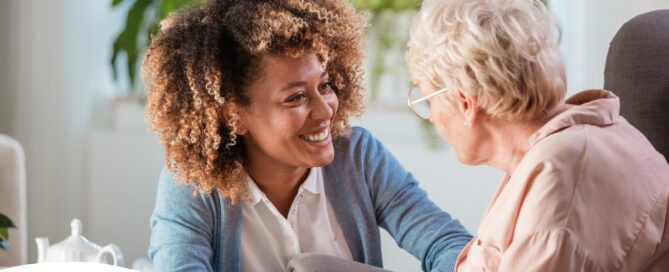 A caregiver smiles at an older client in a chair, representing the joy that can come from a healthy work-life balance.