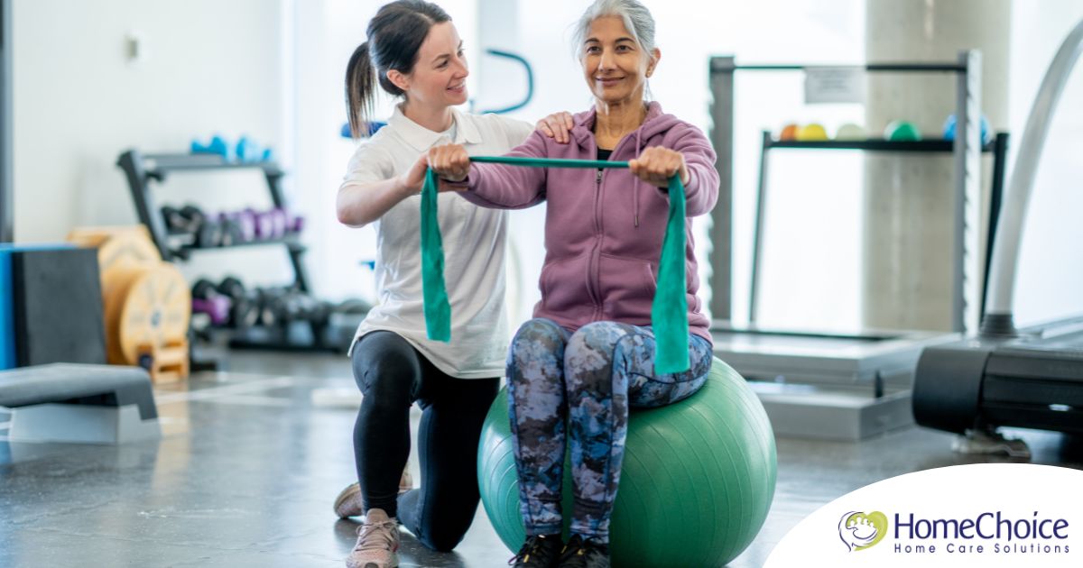 A care provider helps an older woman exercise with a resistance band and an exercise ball, representing how exercise can help with senior fall prevention.