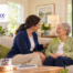An older woman smiles while talking with a respite caregiver as her adult daughter leaves the home feeling reassured.