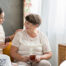 Smiling caregiver with her hand on an older woman’s shoulder as they share tea on the couch, showing home care vs assisted living