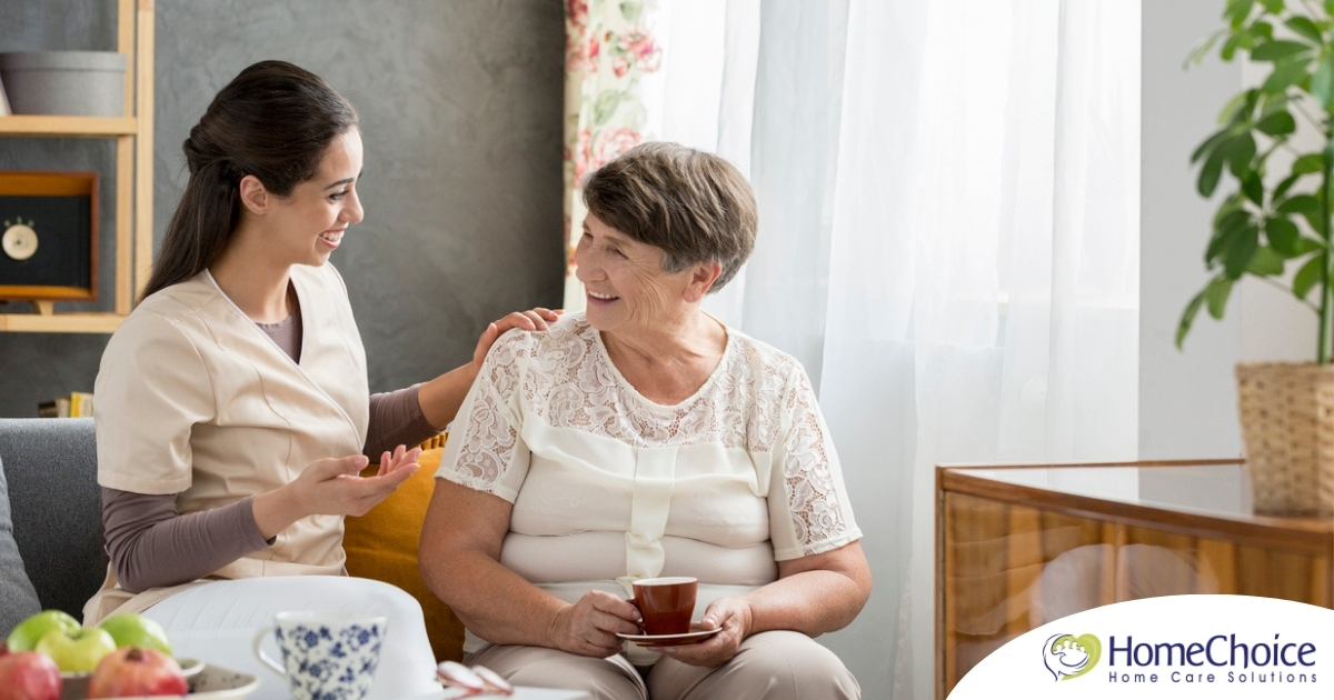 Smiling caregiver with her hand on an older woman’s shoulder as they share tea on the couch, showing home care vs assisted living