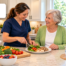 HomeChoice caregiver in navy scrubs and an older woman smiling while preparing a balanced meal with fresh vegetables, fruit and water in a bright home kitchen.